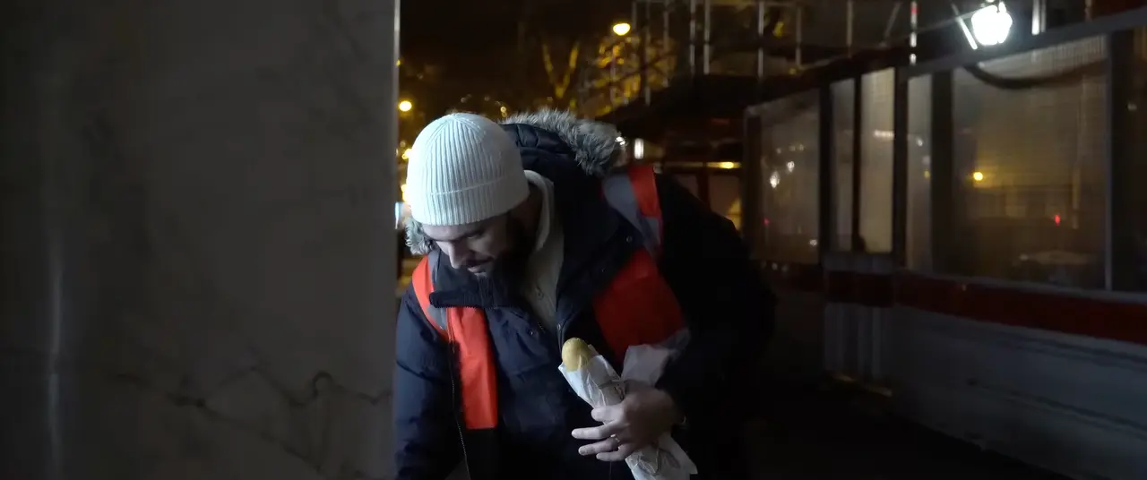 La nuit, dans une rue éclairée, un bénévole au bonnet blanc se penche, une baguette sous le bras.