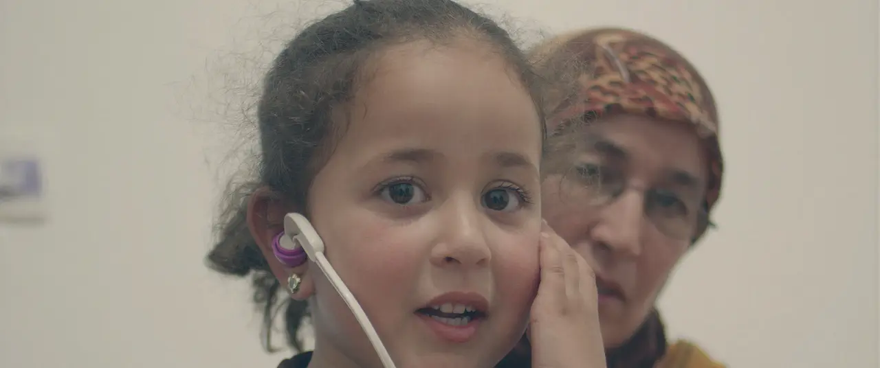 Une petite fille, un stéthoscope jouet contre l’oreille, et derrière elle une femme au foulard.
