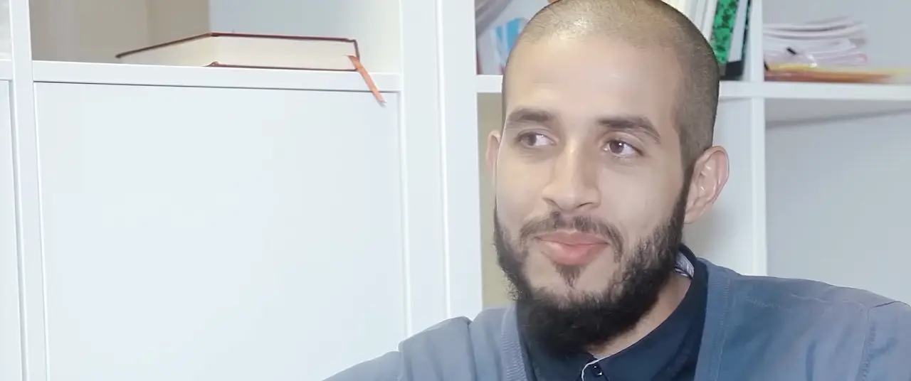 Un homme barbu, assis dans un bureau aux étagères blanches, pendant un entretien.