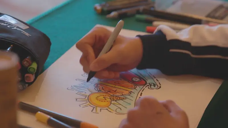 Des mains dessinent au feutre sur une feuille, entourées de crayons.