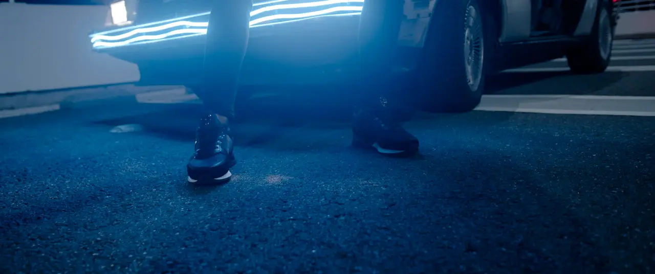 Des baskets noires sur l’asphalte, devant une voiture éclairée de néons bleus.